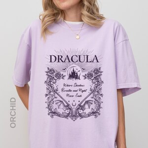 May include: A light purple t-shirt featuring a dark purple graphic print. The design includes the text "DRACULA" above a castle silhouette, bats, roses, and the text "Where Shadows Breathe and Night Never Ends" framed by ornate scrollwork.