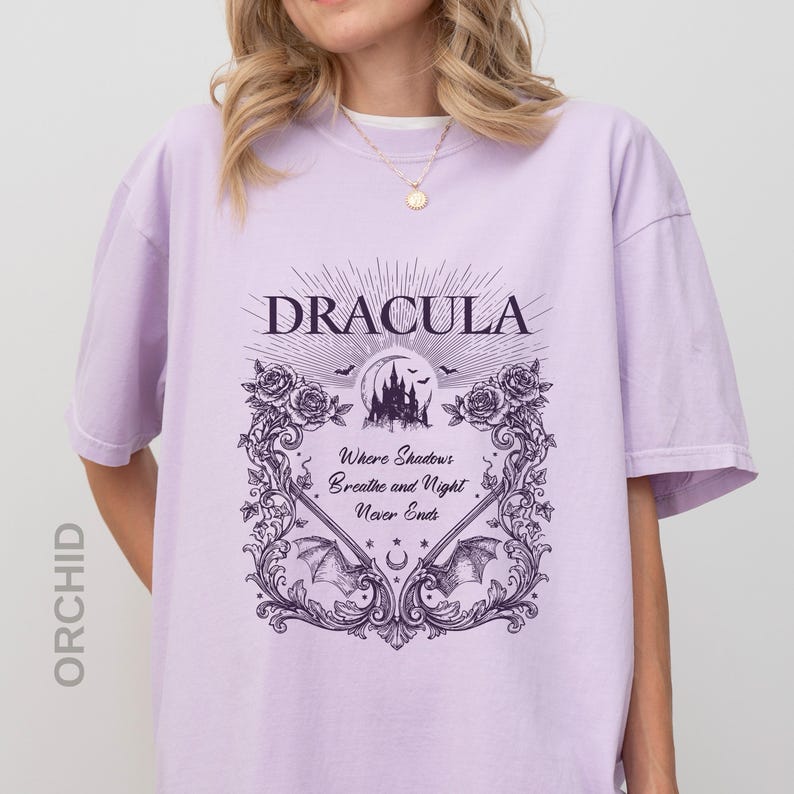 May include: A light purple t-shirt featuring a dark purple graphic print. The design includes the text "DRACULA" above a castle silhouette, bats, roses, and the text "Where Shadows Breathe and Night Never Ends" framed by ornate scrollwork.