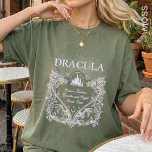 May include: An olive green t-shirt featuring a white graphic print of a castle, roses, bats, and the text "DRACULA" above the phrase "Where Shadows Breathe and Night Never Ends" enclosed in an ornate, decorative rectangular frame.