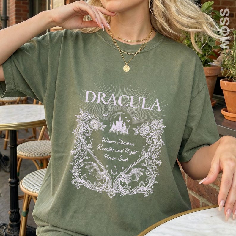 May include: An olive green t-shirt featuring a white graphic print of a castle, roses, bats, and the text "DRACULA" above the phrase "Where Shadows Breathe and Night Never Ends" enclosed in an ornate, decorative rectangular frame.