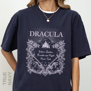 May include: A true navy short-sleeve t-shirt featuring a light purple graphic print. The design includes the text "DRACULA" and "Where Shadows Breathe and Night Never Ends," surrounded by illustrations of a castle, roses, bats, and vines.