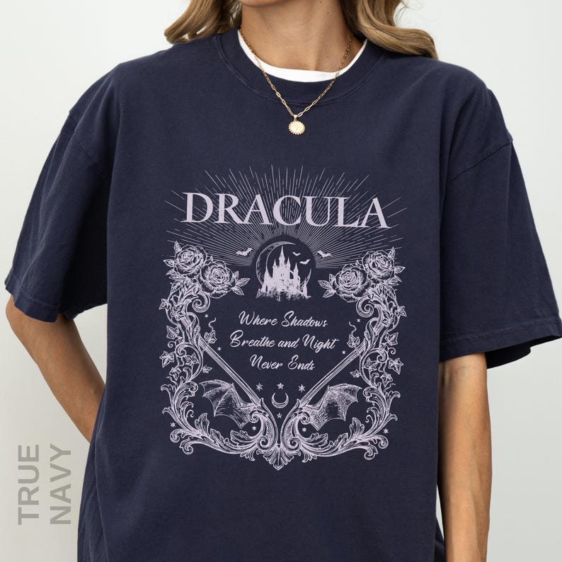 May include: A true navy short-sleeve t-shirt featuring a light purple graphic print. The design includes the text "DRACULA" and "Where Shadows Breathe and Night Never Ends," surrounded by illustrations of a castle, roses, bats, and vines.