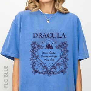 May include: A blue short-sleeved t-shirt featuring a dark purple graphic print. The design includes the text "DRACULA" above a castle, bats, and roses, with the phrase "Where Shadows Breathe and Night Never Ends" centered within an ornate frame.