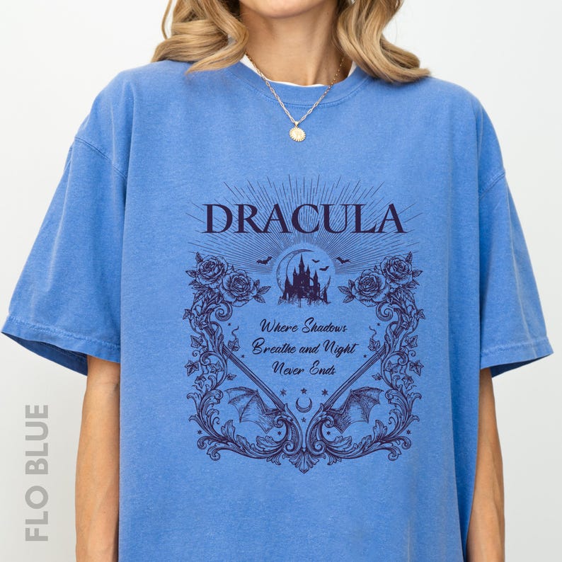 May include: A blue short-sleeved t-shirt featuring a dark purple graphic print. The design includes the text "DRACULA" above a castle, bats, and roses, with the phrase "Where Shadows Breathe and Night Never Ends" centered within an ornate frame.