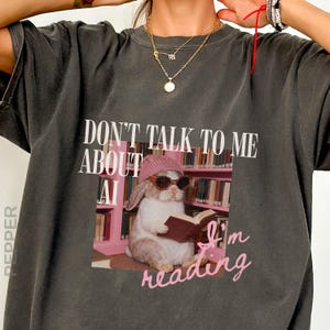 May include: A charcoal gray short-sleeve t-shirt featuring a graphic of a rabbit wearing a pink beanie and sunglasses while reading a book in a library. The text reads "DON'T TALK TO ME ABOUT AI" in white and "I'm reading" in pink script.