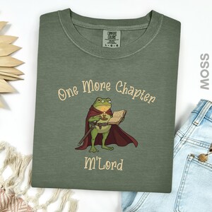 May include: Moss green t-shirt with the text "One More Chapter" and "M'Lord". The graphic features a frog in a cape holding a candle and a book. The shirt is a Comfort Colors brand, size L-G-G.