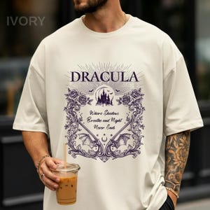 May include: An ivory t-shirt featuring a dark purple graphic print. The design includes the text "DRACULA" above a castle silhouette, bats, and roses. Below, the text reads "Where Shadows Breathe and Night Never Ends" framed by ornate scrollwork.