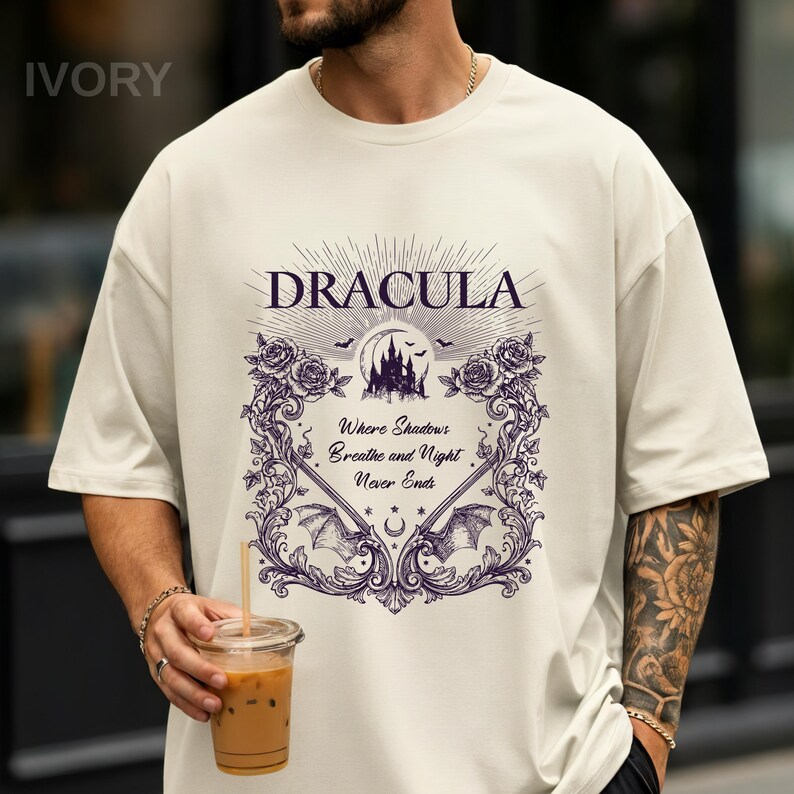May include: An ivory t-shirt featuring a dark purple graphic print. The design includes the text "DRACULA" above a castle silhouette, bats, and roses. Below, the text reads "Where Shadows Breathe and Night Never Ends" framed by ornate scrollwork.