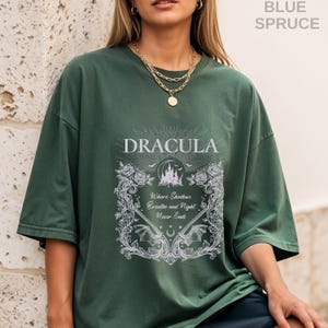 May include: A dark green short-sleeve t-shirt featuring a white graphic print. The design includes the text "DRACULA" and "Where Shadows Breathe and Night Never Ends," surrounded by ornate floral borders, bat wings, and a castle silhouette.