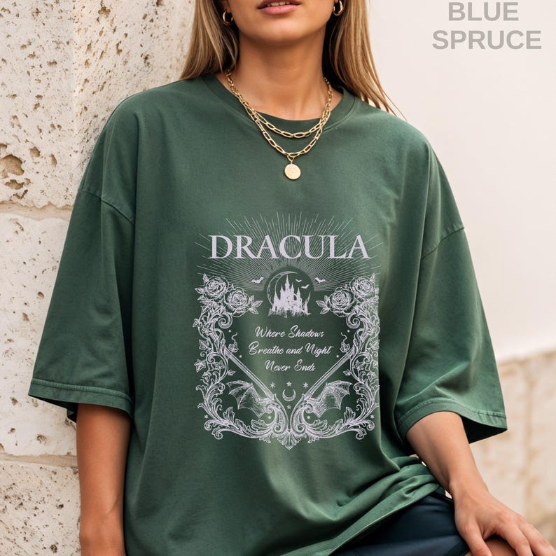 May include: A dark green short-sleeve t-shirt featuring a white graphic print. The design includes the text "DRACULA" and "Where Shadows Breathe and Night Never Ends," surrounded by ornate floral borders, bat wings, and a castle silhouette.