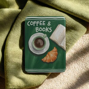 Könnte beinhalten: Ein grünes, buchförmiges Objekt mit den Worten "COFFEE & BOOKS" in Weiß. Das Cover zeigt eine Illustration einer Kaffeetasse, Untertasse, eines offenen Buches und eines Croissants. Weiße Linien umranden die Ränder des Buches.