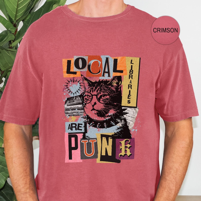 May include: A crimson short-sleeve t-shirt featuring a graphic print of a cat wearing glasses and a spiked collar. The text reads LOCAL LIBRARIES ARE PUNK. The design includes collage-style elements with orange, blue, and black accents.