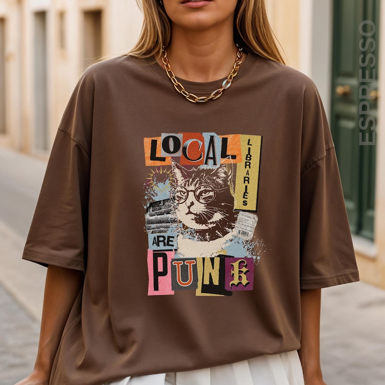 May include: A brown short-sleeve t-shirt featuring a graphic print of a cat wearing glasses. The text reads LOCAL LIBRARIES ARE PUNK. The design includes collage-style elements in orange, blue, and yellow, with a thick gold chain necklace.