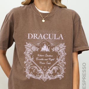 May include: A brown short-sleeve t-shirt featuring a light purple graphic print. The design includes the text DRACULA, a castle, bats, roses, and the phrase Where Shadows Breathe and Night Never Ends, framed by ornate scrollwork and vines.