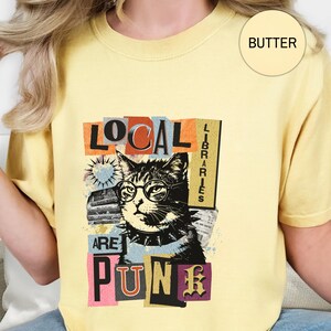 May include: A butter yellow t-shirt featuring a graphic of a cat wearing round glasses and a spiked collar. The text reads "LOCAL LIBRARIES ARE PUNK" in a collage style with various fonts and colours. A small circle in the corner says "BUTTER".
