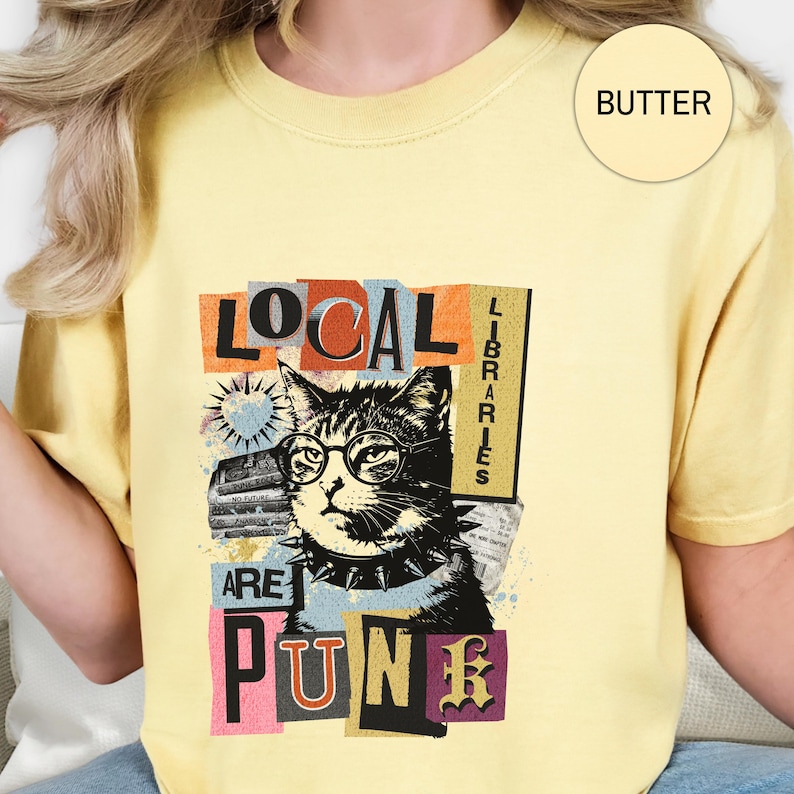May include: A butter yellow t-shirt featuring a graphic of a cat wearing round glasses and a spiked collar. The text reads "LOCAL LIBRARIES ARE PUNK" in a collage style with various fonts and colours. A small circle in the corner says "BUTTER".