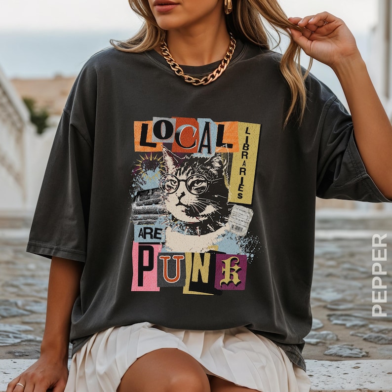 May include: A charcoal grey short-sleeve t-shirt featuring a graphic print of a cat wearing round glasses. The text reads "LOCAL LIBRARIES ARE PUNK" in a collage style with orange, yellow, blue, and pink accents. The design includes a library card.