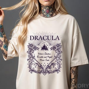 May include: An ivory t-shirt featuring a dark purple graphic print. The design includes the text "DRACULA" above a castle silhouette, moon, and bats. Below, the text reads "Where Shadows Breathe and Night Never Ends" framed by roses and wings.