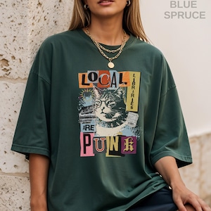 May include: A blue spruce coloured short-sleeve t-shirt featuring a graphic print of a cat wearing glasses. The text reads "LOCAL LIBRARIES ARE PUNK" in a collage style with various colours. The shirt has a crew neckline and a loose, relaxed fit.