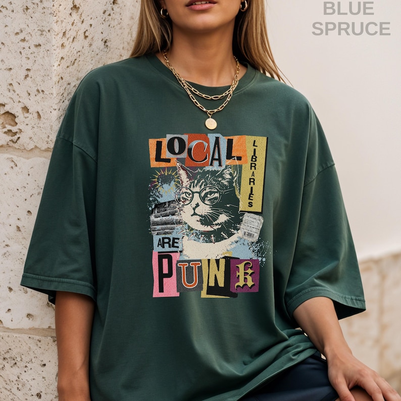 May include: A blue spruce coloured short-sleeve t-shirt featuring a graphic print of a cat wearing glasses. The text reads "LOCAL LIBRARIES ARE PUNK" in a collage style with various colours. The shirt has a crew neckline and a loose, relaxed fit.