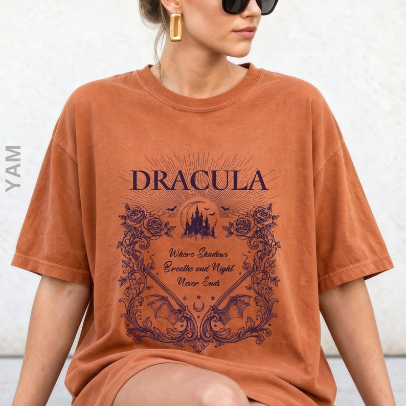May include: A burnt orange short-sleeve t-shirt featuring a dark purple graphic print. The design includes the text "DRACULA" above a castle silhouette, roses, bats, and the text "Where Shadows Breathe and Night Never Ends" in a frame.