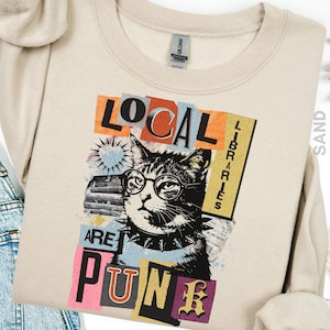 May include: A sand-colored sweatshirt featuring a graphic of a cat wearing glasses and a studded collar. The design includes the text "LOCAL LIBRARIES ARE PUNK" in various colors and fonts, with a collage-style background.