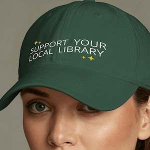 Peut inclure: Casquette de baseball vert foncé avec l'inscription blanche "SUPPORT YOUR LOCAL LIBRARY", ornée d'étoiles jaunes. La casquette a une visière incurvée et une conception classique et réglable.