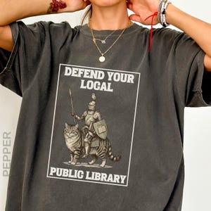 May include: A charcoal gray t-shirt featuring a graphic print of a knight in armor riding a large tabby cat. The knight holds a spear and a shield with a book icon. The text reads DEFEND YOUR LOCAL PUBLIC LIBRARY in white capital letters.
