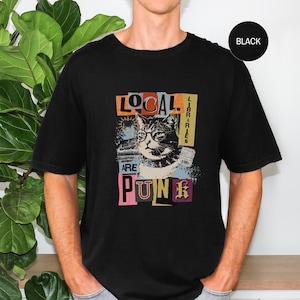 May include: A black short-sleeved t-shirt featuring a graphic print of a cat wearing round glasses. The text reads LOCAL LIBRARIES ARE PUNK, arranged in a collage style with various fonts and colours including orange, white, and purple accents.