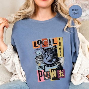May include: A blue jean coloured t-shirt featuring a graphic print of a cat wearing glasses and a spiked collar. The text reads LOCAL LIBRARIES ARE PUNK. The design includes colourful rectangular blocks and a small heart shape behind the cat's head.