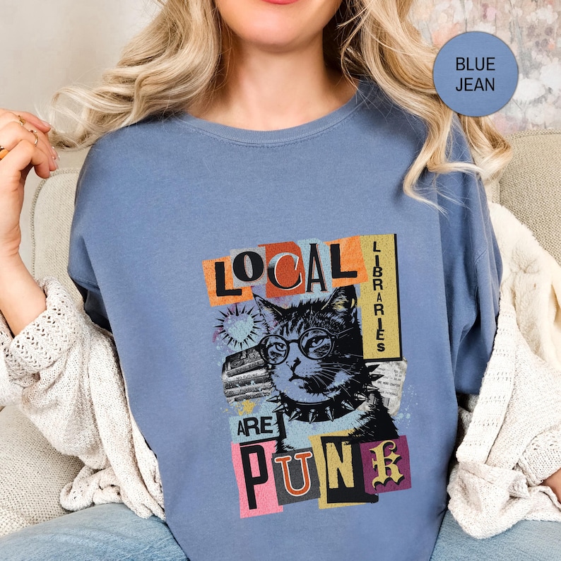 May include: A blue jean coloured t-shirt featuring a graphic print of a cat wearing glasses and a spiked collar. The text reads LOCAL LIBRARIES ARE PUNK. The design includes colourful rectangular blocks and a small heart shape behind the cat's head.