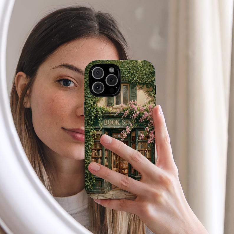 K&ouml;nnte beinhalten: Eine Smartphone-H&uuml;lle mit einer aufgedruckten Illustration einer Buchhandlung mit gr&uuml;nem Efeu an den W&auml;nden, rosa Blumen und dem Schriftzug BOOK SHOP &uuml;ber dem Eingang, wobei B&uuml;cherregale durch die Glasfenster zu sehen sind.