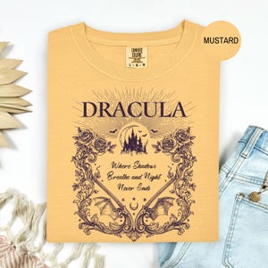 May include: A mustard yellow t-shirt featuring a dark purple graphic print. The design includes the text "DRACULA" above a castle silhouette, bats, and the phrase "Where Shadows Breathe and Night Never Ends" framed by ornate floral vines.