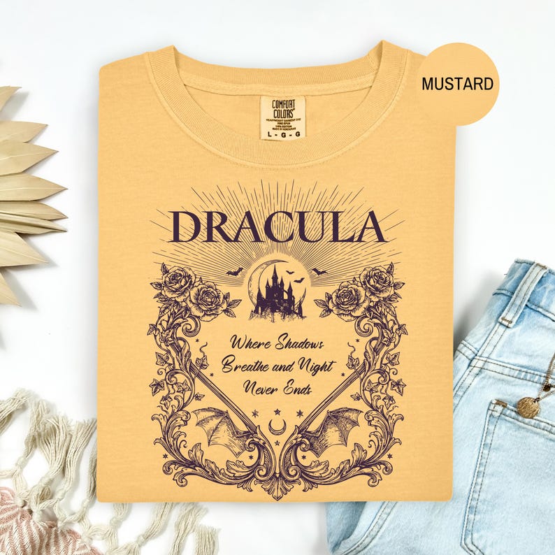 May include: A mustard yellow t-shirt featuring a dark purple graphic print. The design includes the text "DRACULA" above a castle silhouette, bats, and the phrase "Where Shadows Breathe and Night Never Ends" framed by ornate floral vines.