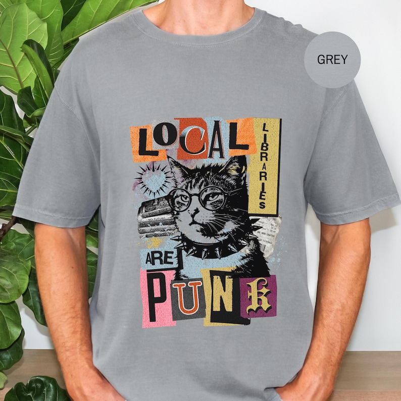 May include: A grey short-sleeve t-shirt featuring a graphic of a cat wearing round glasses and a spiked collar. The text reads "LOCAL LIBRARIES ARE PUNK" in a collage-style layout with colourful rectangular accents and a small heart icon.