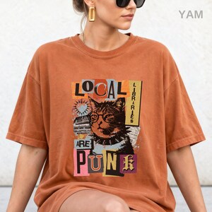May include: A rust-coloured short-sleeve t-shirt featuring a graphic print of a cat wearing round glasses and a spiked collar. The text reads LOCAL LIBRARIES ARE PUNK, arranged in a collage style with various rectangular shapes and colours.