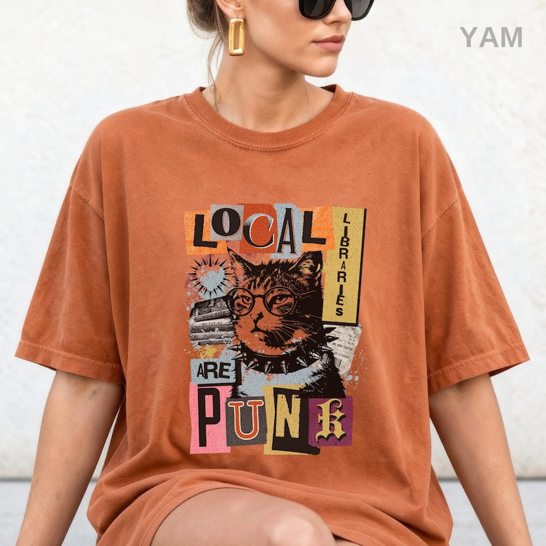 May include: A rust-coloured short-sleeve t-shirt featuring a graphic print of a cat wearing round glasses and a spiked collar. The text reads LOCAL LIBRARIES ARE PUNK, arranged in a collage style with various rectangular shapes and colours.