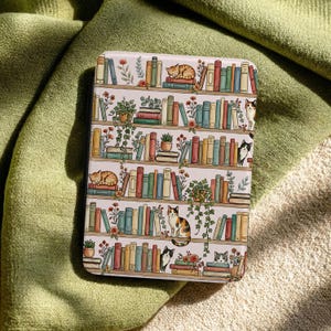 May include: A rectangular case featuring a bookshelf design with cats. The illustration shows rows of books in various colors, potted plants, and sleeping cats. The case is set against a green and beige fabric background.