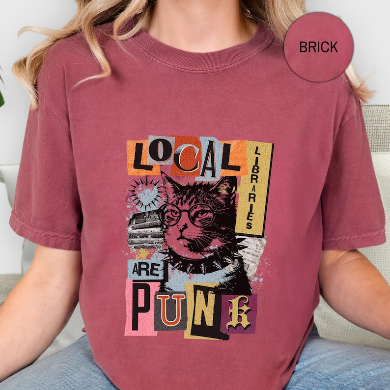 May include: A brick-coloured short-sleeve t-shirt featuring a graphic print of a cat wearing round glasses and a spiked collar. The text reads LOCAL LIBRARIES ARE PUNK. The design includes collage-style elements, a heart, and star shapes.