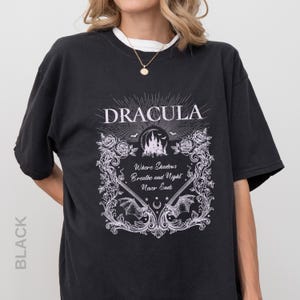 May include: A black t-shirt featuring a white graphic print with the text "DRACULA" and "Where Shadows Breathe and Night Never Ends." The design includes a castle silhouette, bats, roses, and ornate scrollwork framing the central text.