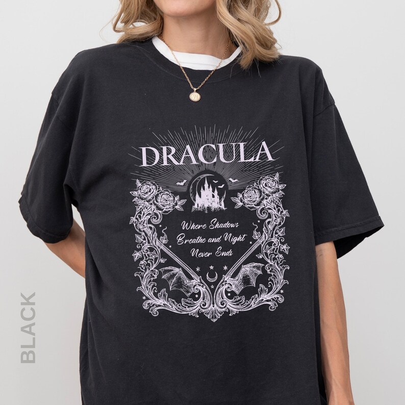 May include: A black t-shirt featuring a white graphic print with the text "DRACULA" and "Where Shadows Breathe and Night Never Ends." The design includes a castle silhouette, bats, roses, and ornate scrollwork framing the central text.