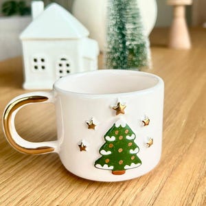 Op de afbeelding: Een witte keramische mok met een gouden oor en rand, versierd met een groene kerstboom en gouden sterren. De mok staat op een houten oppervlak, met een wit huis en andere feestelijke decoraties op de achtergrond.