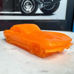 May include: An orange toy car with a classic design. The car is a coupe with visible wheels and a detailed front end. The toy car is sitting on a white surface, with a framed image of a vintage car in the background.