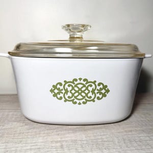 May include: White casserole dish with a clear glass lid and a clear glass knob. The dish has a green decorative design on the front. The dish has two small handles on either side.