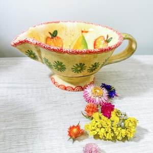May include: A yellow ceramic gravy boat with a red and green floral design. The interior features painted fruit, and the rim is trimmed with red. A green handle is on the side. Dried flowers are scattered around the base.