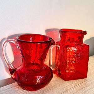 May include: Two vibrant red glass pitchers with a textured, crinkled surface. The left pitcher features a rounded body and a wide, flared rim, while the right pitcher has a square-shaped base and body. Both vessels include clear glass handles.