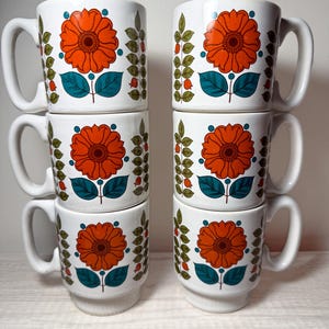 May include: Six white ceramic mugs with a retro floral design. Each mug features a large orange flower with a teal center and teal leaves, along with green and red accents. The mugs have a classic handle and a slightly flared base.