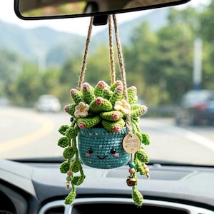 May include: A handmade crochet succulent car charm. The green and pink succulent is in a blue pot with a smiling face. The charm hangs from a rearview mirror with a tag that says "Handmade with Love".