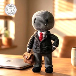 May include: Crocheted doll in a business suit, complete with a red tie and a small briefcase. The doll is gray with black shoes and a white collared shirt. The doll has a round head and is standing on a wooden surface.