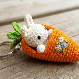 May include: A handmade crochet keychain featuring a white bunny peeking out of an orange carrot. The carrot has green leaves and is decorated with embroidered blue and yellow flowers. The keychain is attached to a silver metal ring.